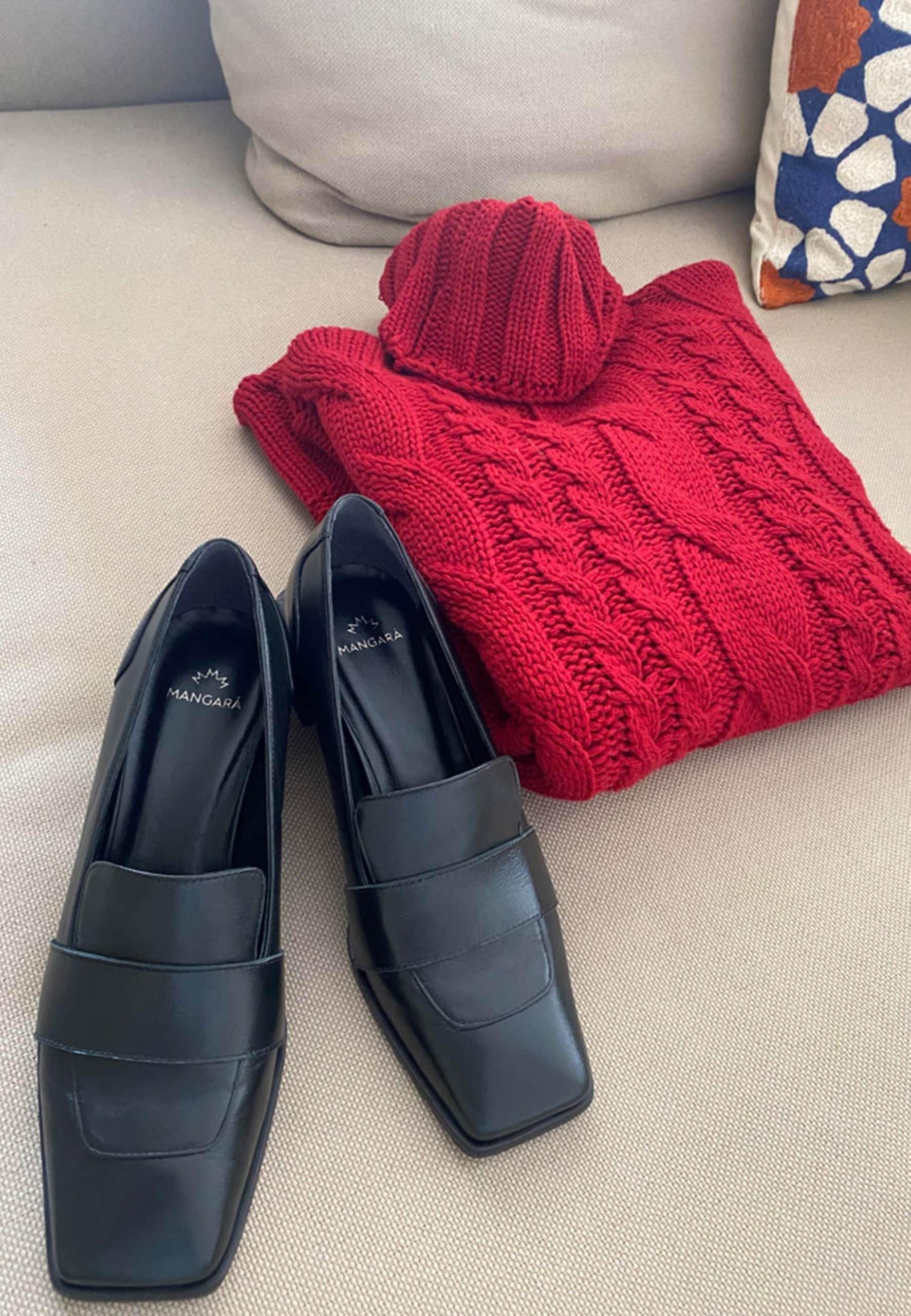 Black loafers on a beige surface with a red knitted sweater and colorful pillow in the background.