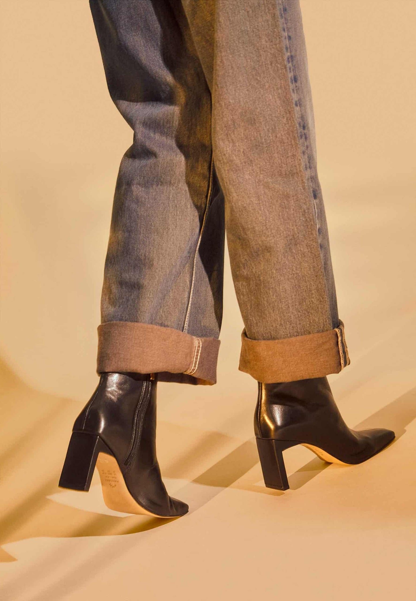 Close-up of feet wearing black high-heeled shoes with rolled-up jeans on a beige background