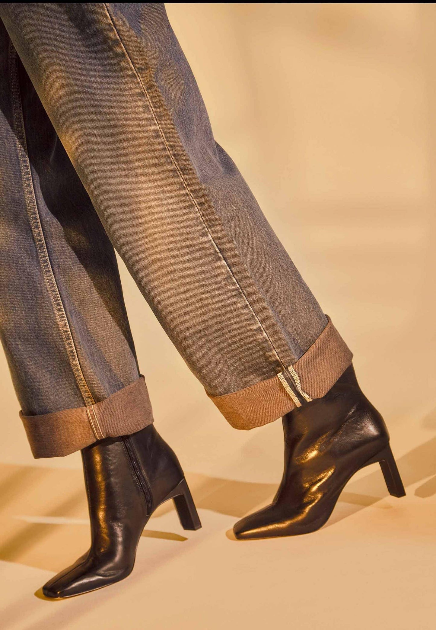 Black ankle boots worn with rolled-up jeans on a beige background