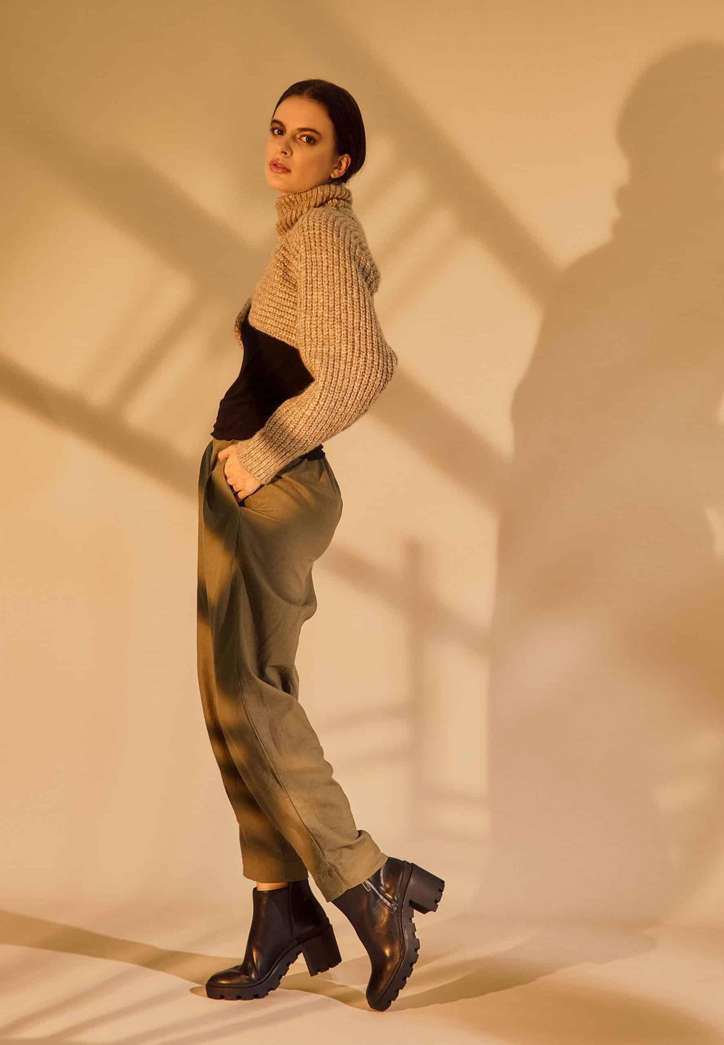 Woman wearing a beige sweater, black top, green pants, and black boots against a plain background