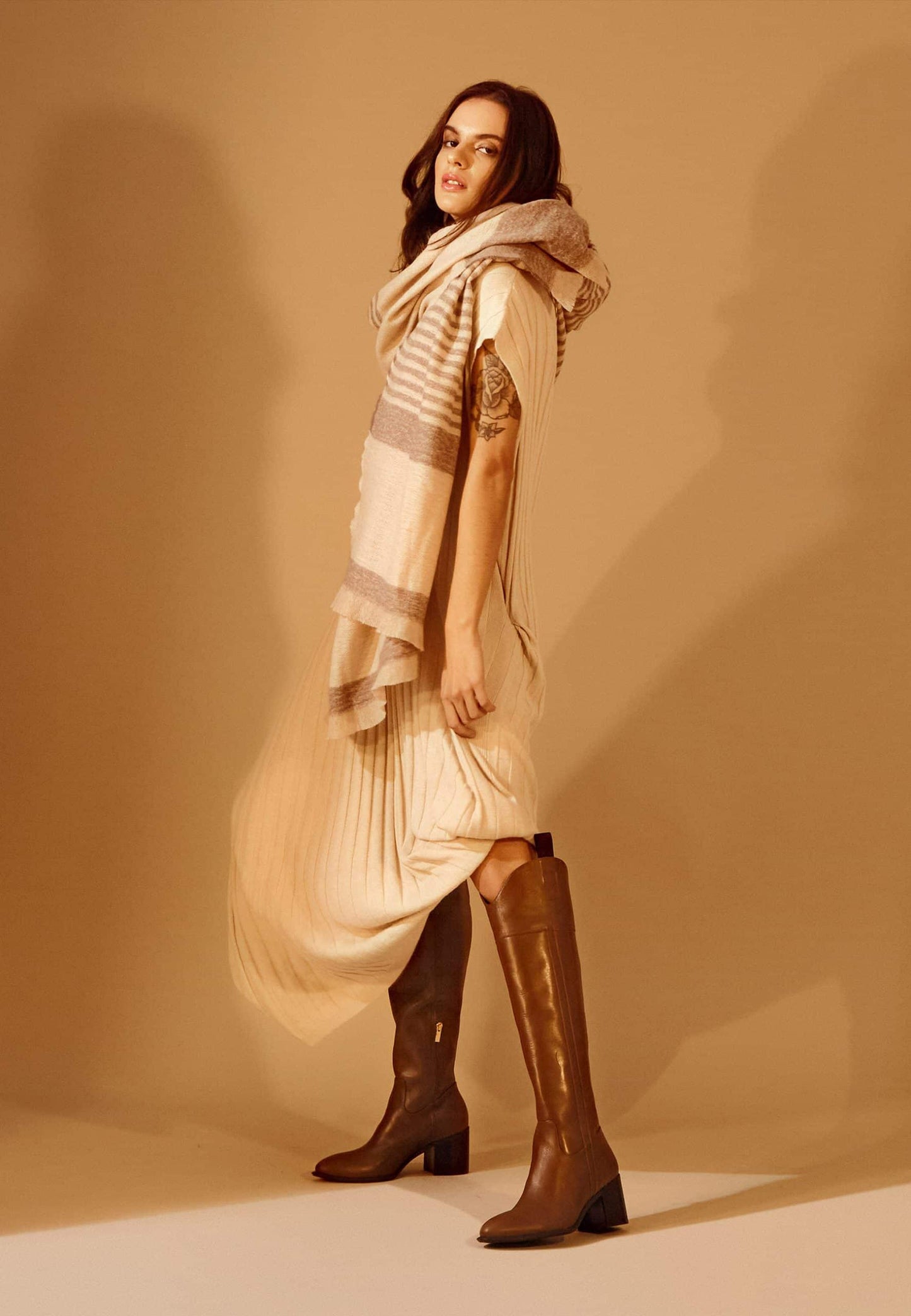 Woman wearing a beige dress, striped scarf, and brown knee-high boots against a beige background