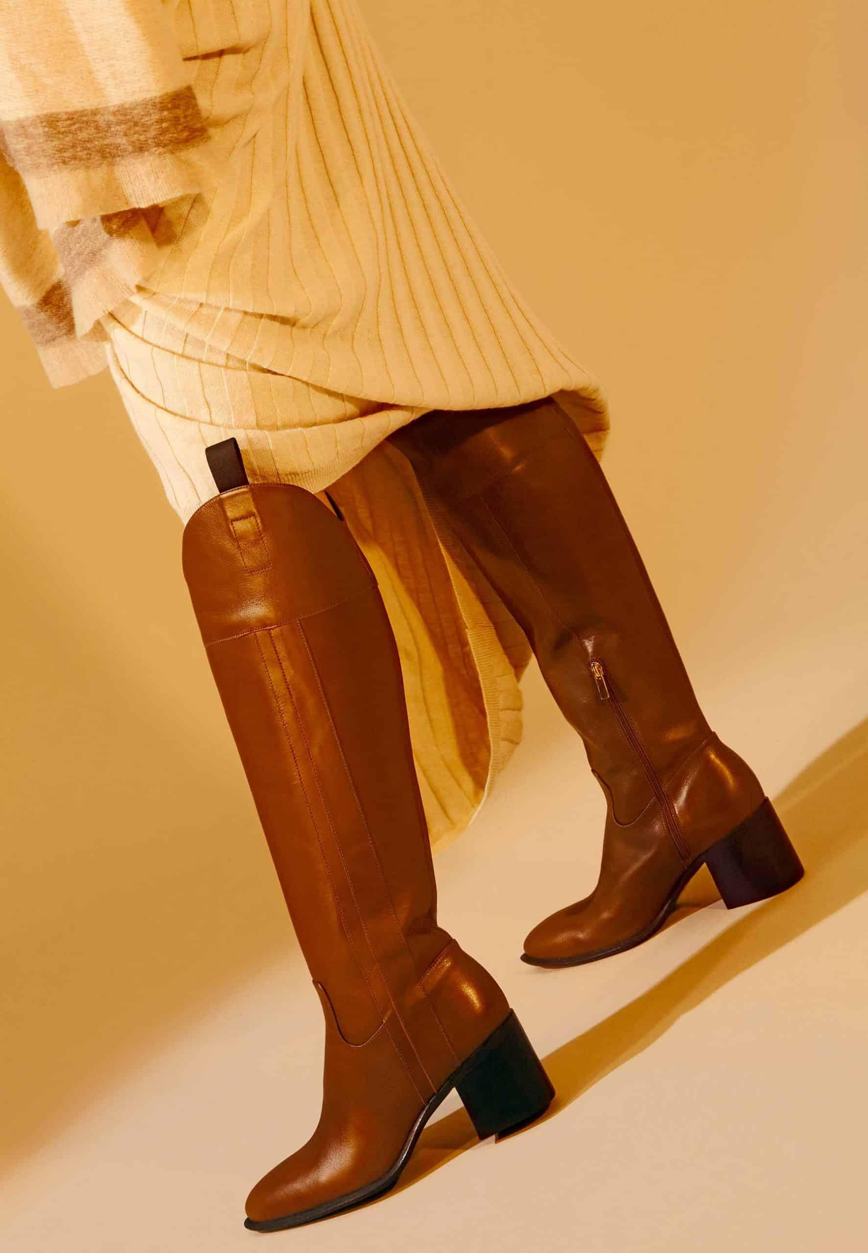 Brown knee-high boots worn with a beige dress on a beige background
