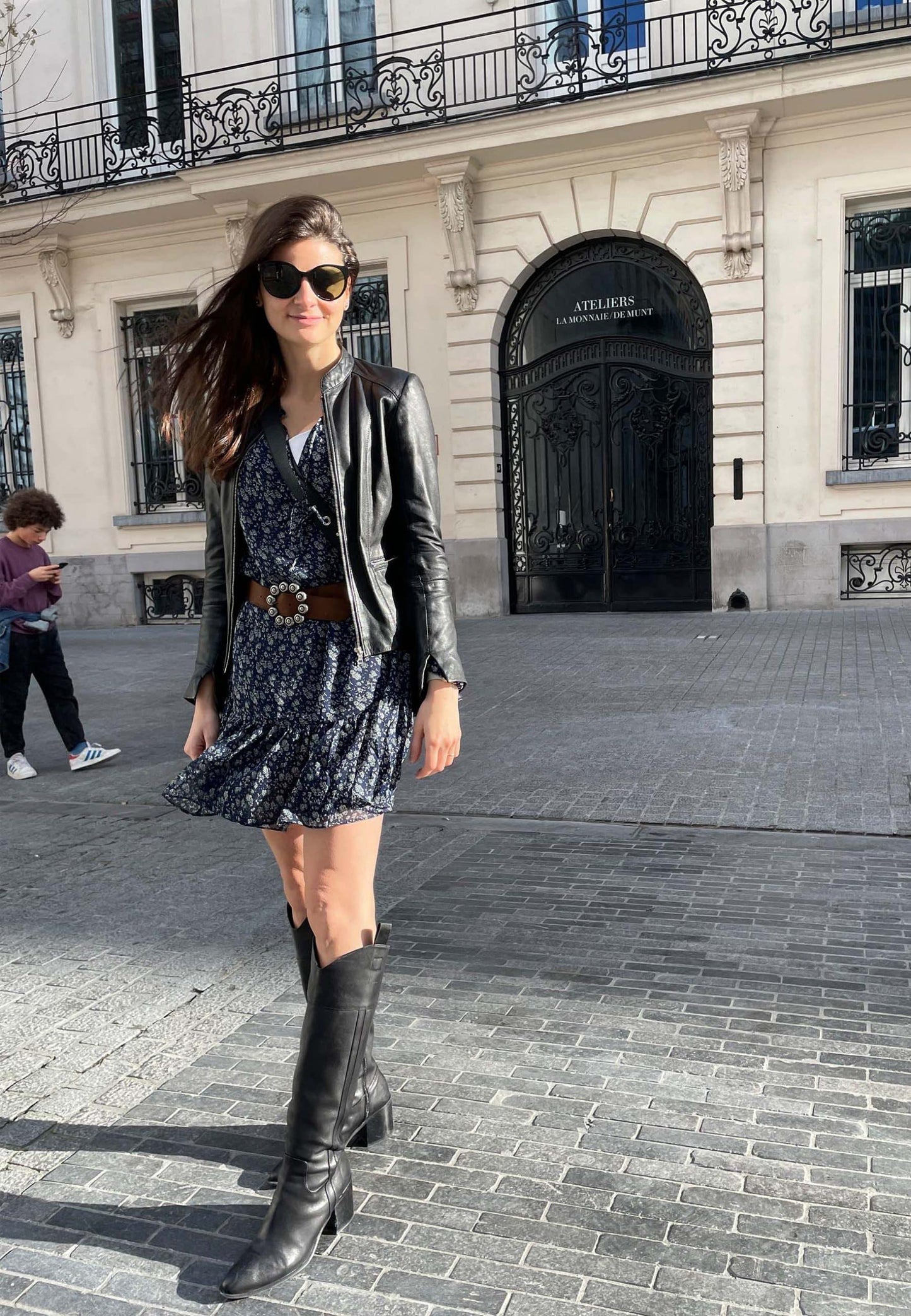 Woman in a black leather jacket and skirt with black boots standing on a street in an urban setting.