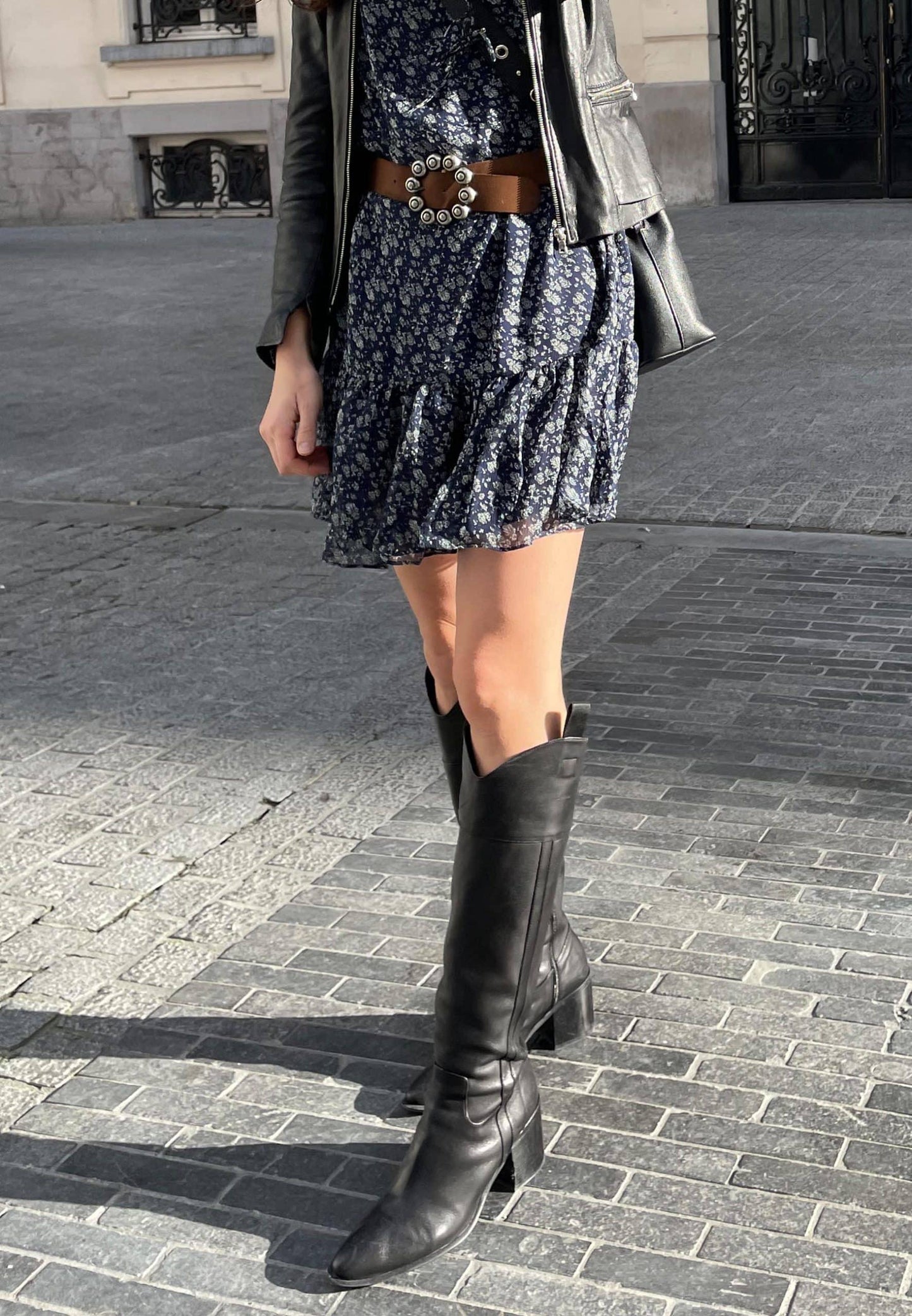 Person wearing a floral dress, leather jacket, and black knee-high boots on a paved street.