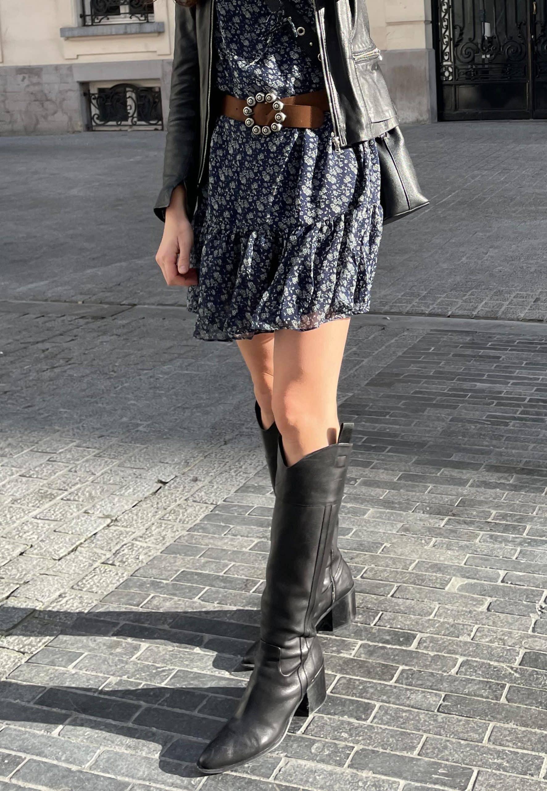 Person wearing a floral dress, leather jacket, and black knee-high boots on a paved street.