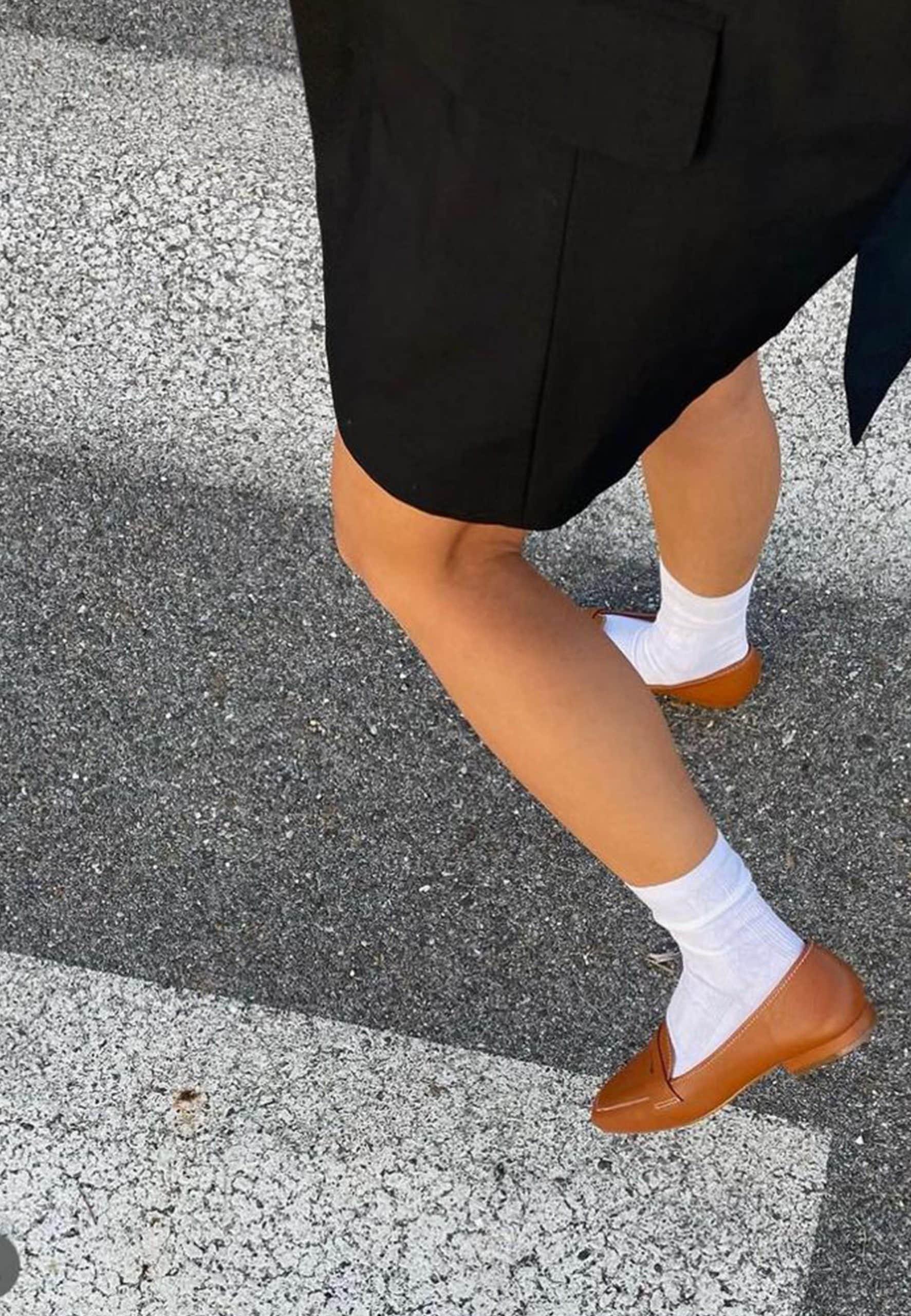 Person wearing a black skirt, white socks, and brown shoes on a road.