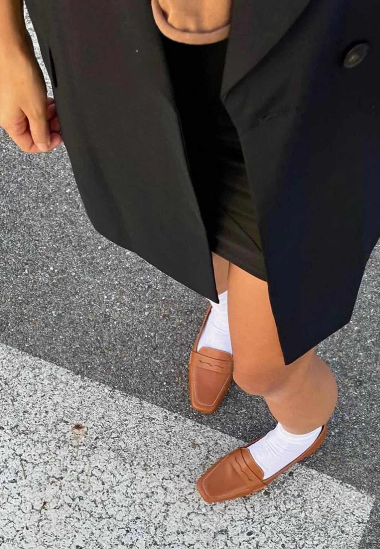 Person wearing a black coat, brown loafers, and white socks on a street.