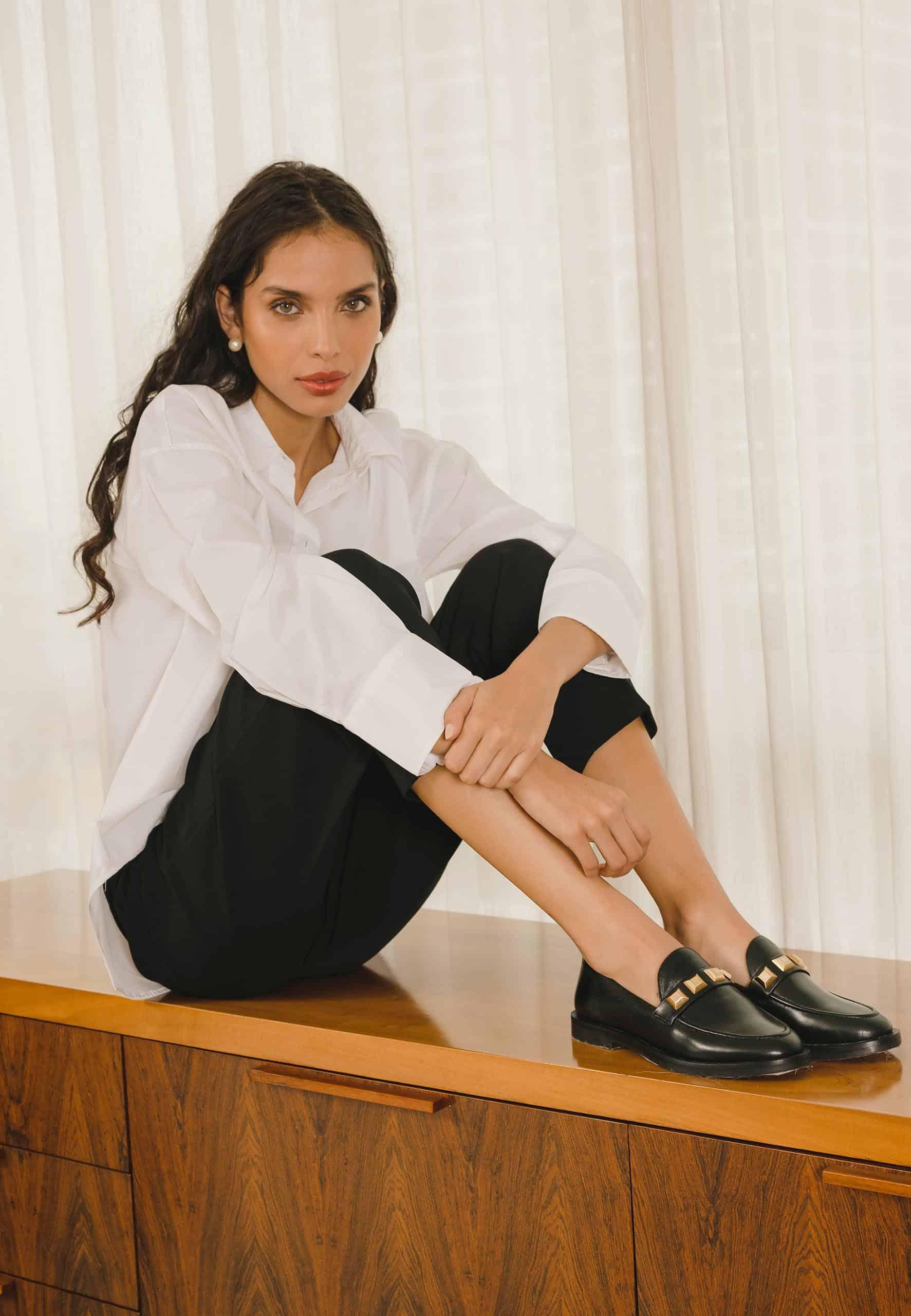 Woman sitting on a wooden surface wearing a white shirt and black pants with gold shoes.