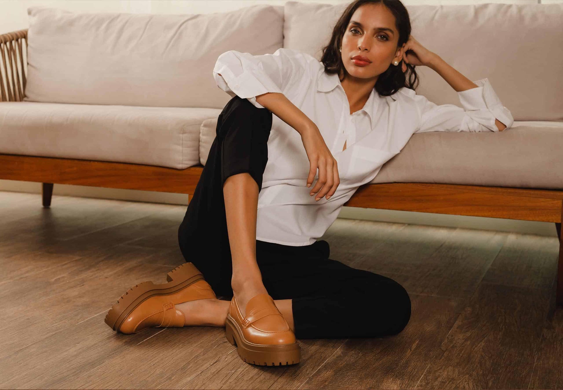 Woman sitting on a couch wearing a white shirt and black pants with brown shoes.