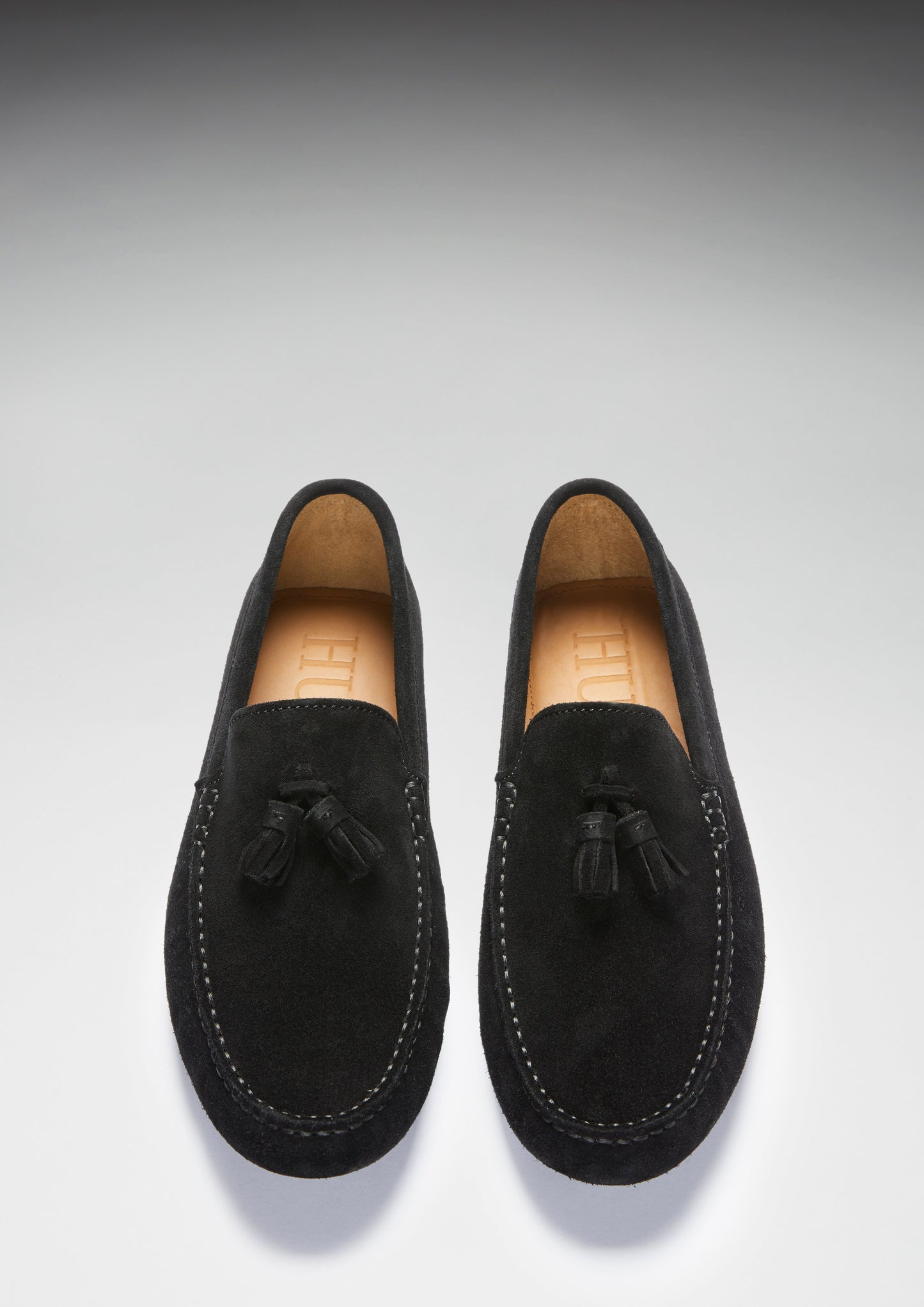Pair of black suede loafers with tassels on a light gray background