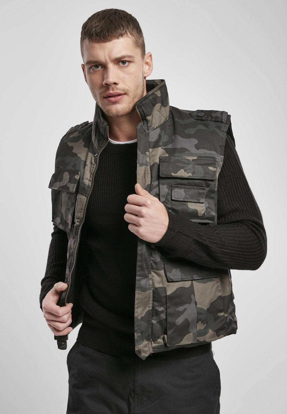 Man wearing a camouflage vest over a black shirt on a light gray background