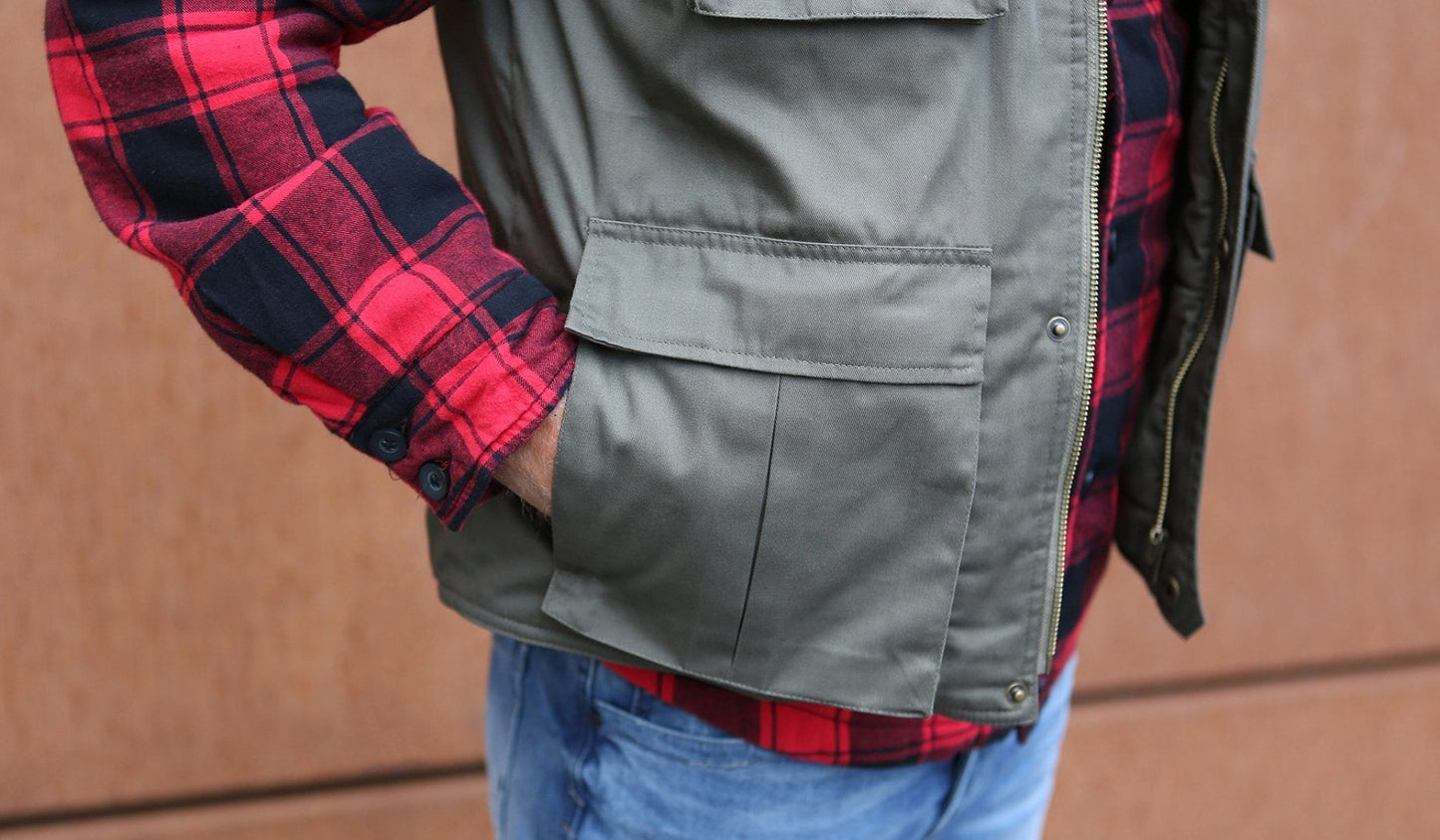 Person wearing a gray jacket over a red and black plaid shirt with blue jeans against a brown background