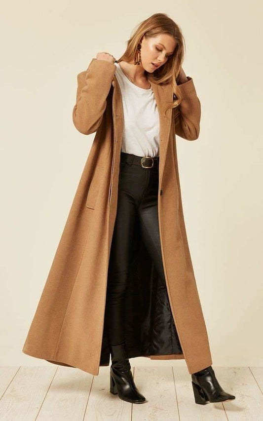 Woman wearing a long brown coat over a white shirt and black pants on a light background