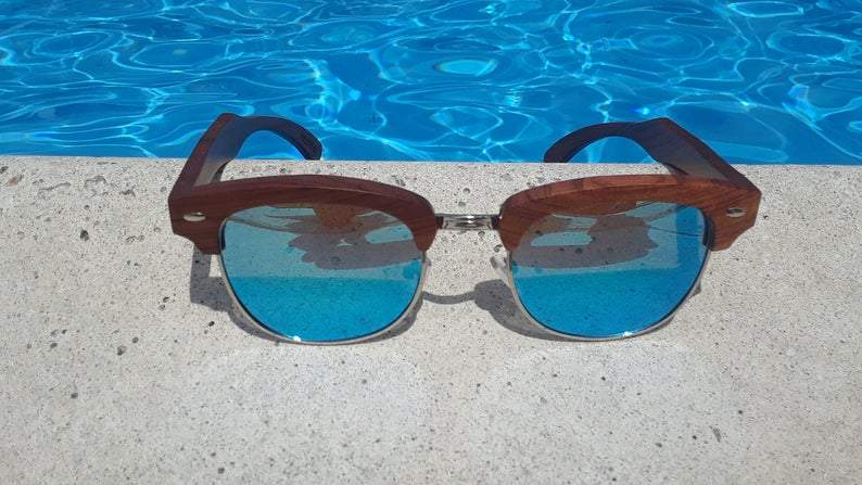 Wooden-framed sunglasses with blue-tinted lenses on a concrete ledge by a pool.