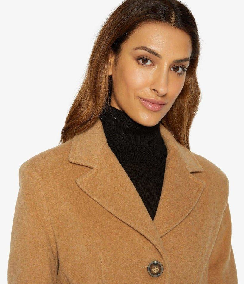 Woman wearing a tan coat over a black turtleneck on a white background