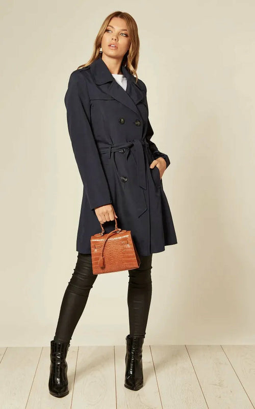 Woman wearing a navy trench coat holding a brown leather bag against a plain background