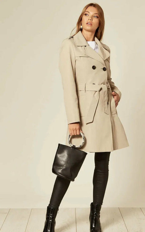 Woman wearing a beige trench coat with a black handbag on a plain background