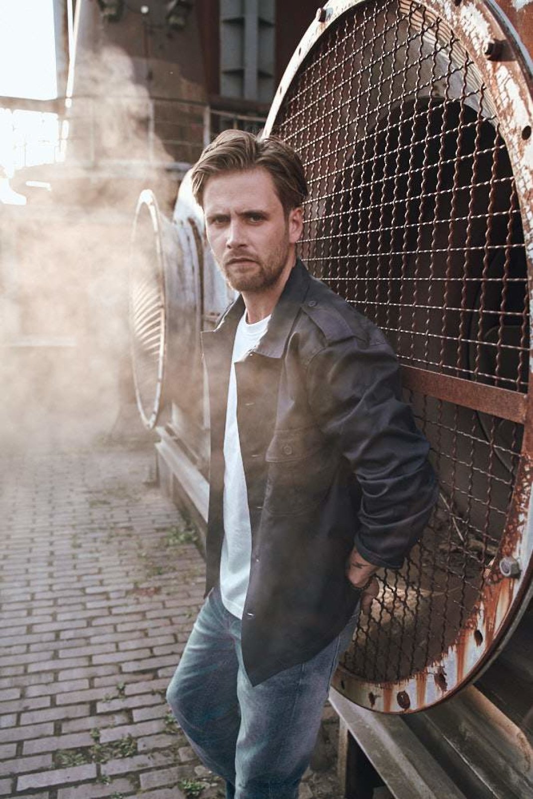 Man leaning against a large metal fan with steam in the background