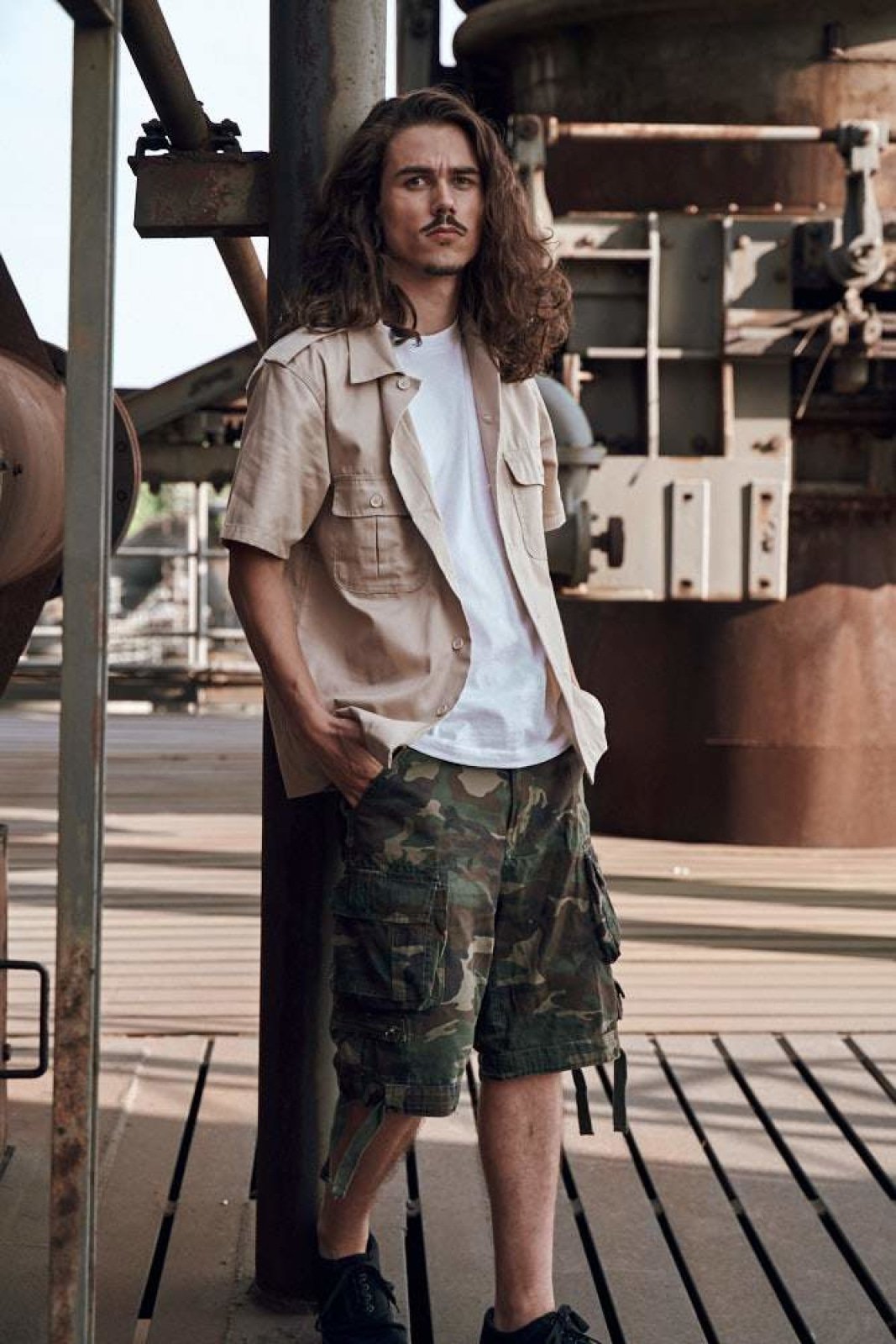 Man wearing a beige shirt, white t-shirt, and camouflage shorts standing in an industrial setting.