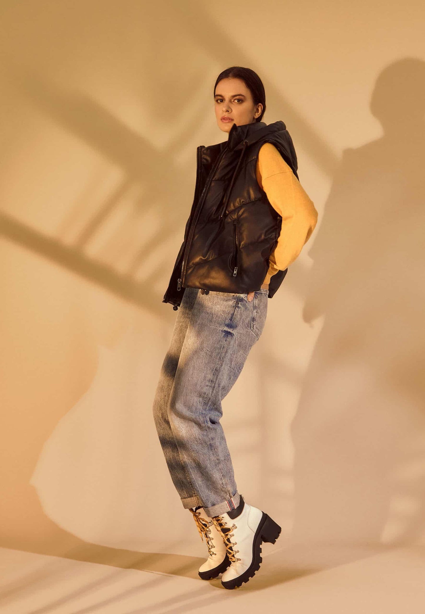 Person wearing a black sleeveless jacket, blue jeans, and white boots against a beige background