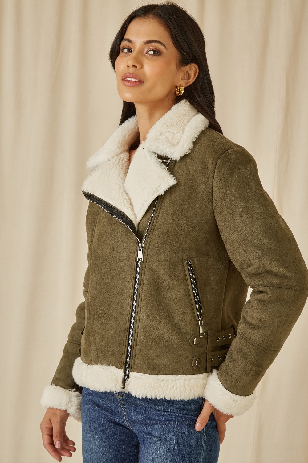 Woman wearing a green suede jacket with white fur lining against a beige background