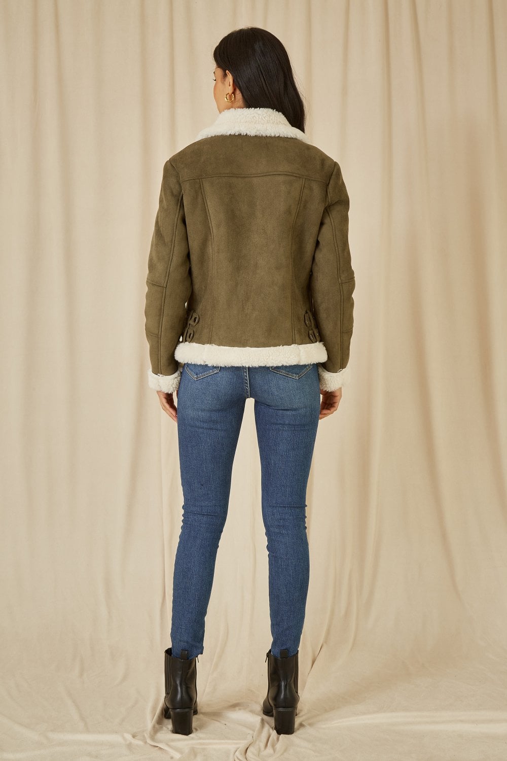 Person wearing a green suede jacket with white fur trim, blue jeans, and black boots on a beige background