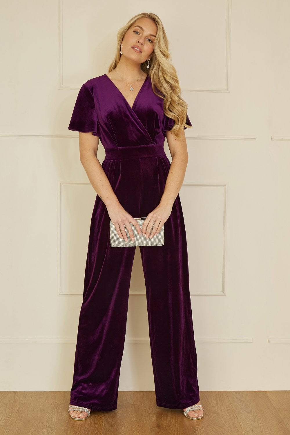 Woman wearing a purple velvet jumpsuit against a white wall.