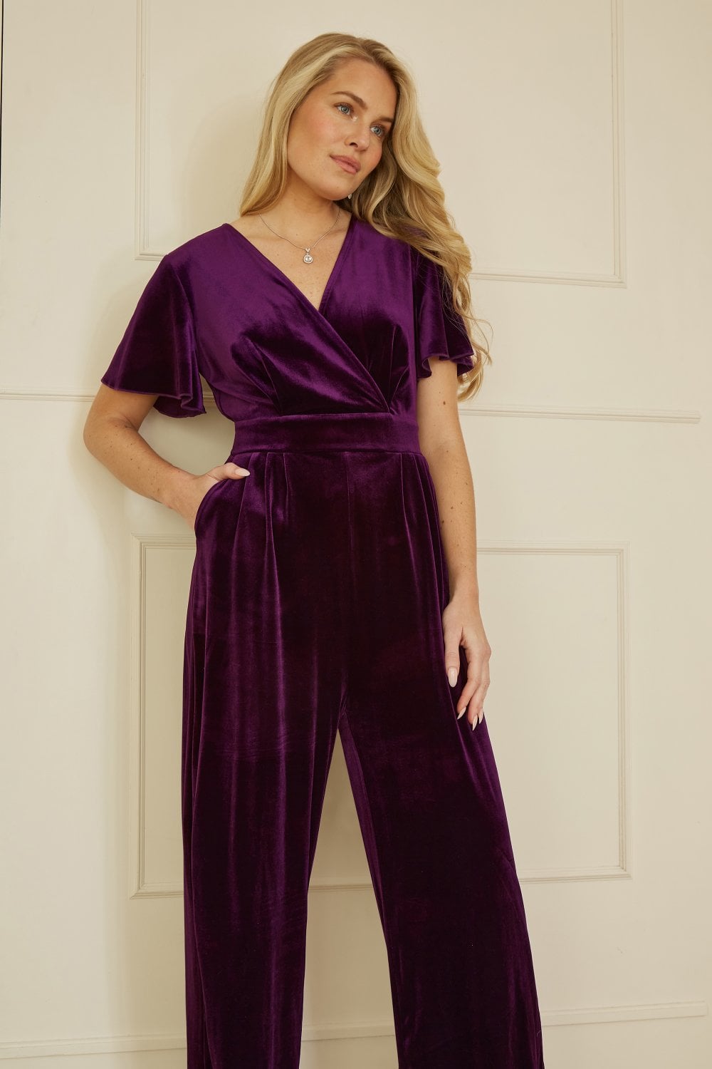 Woman wearing a purple velvet jumpsuit against a white wall.
