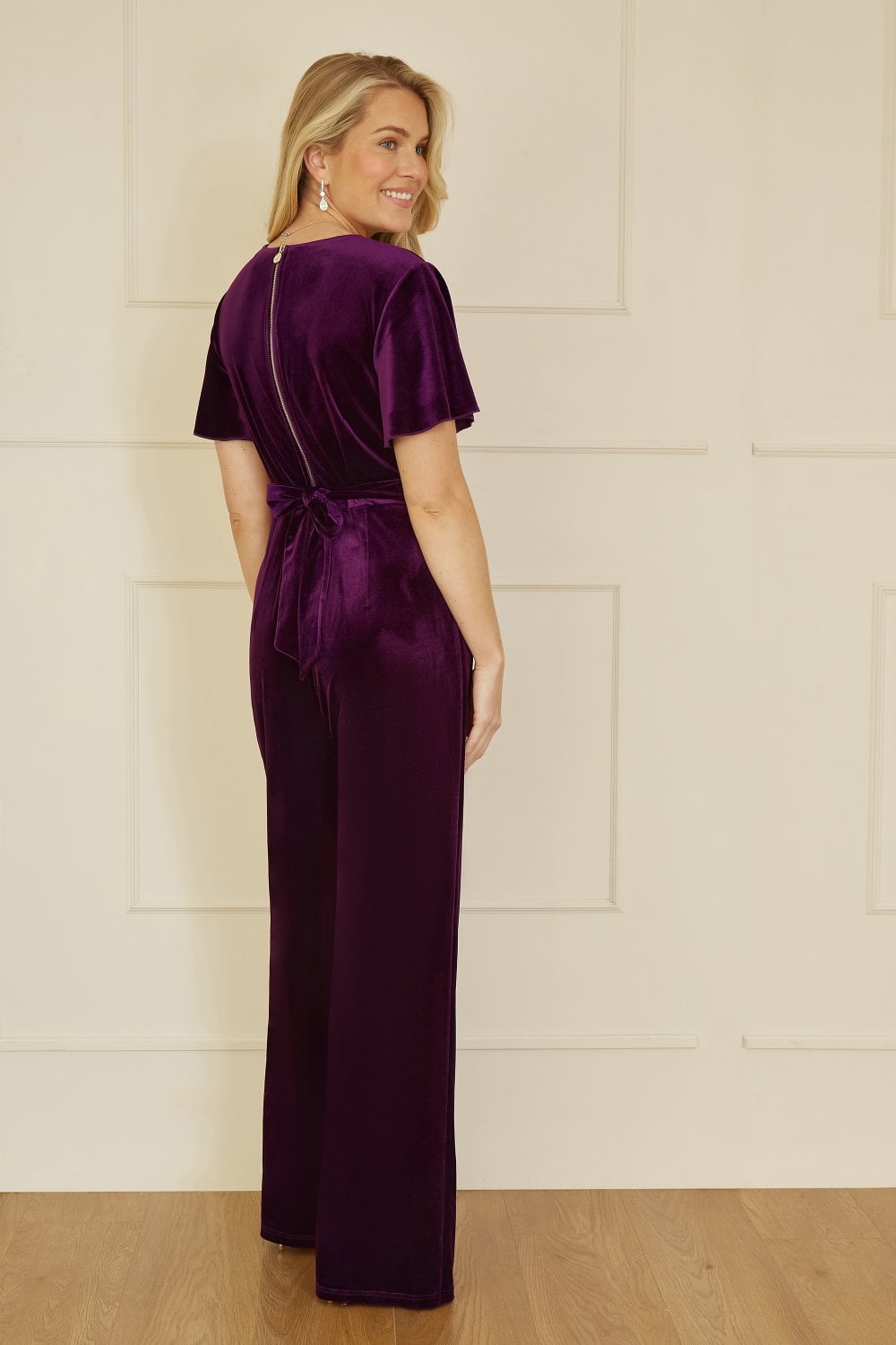 Woman wearing a purple velvet jumpsuit against a light wooden paneled wall.